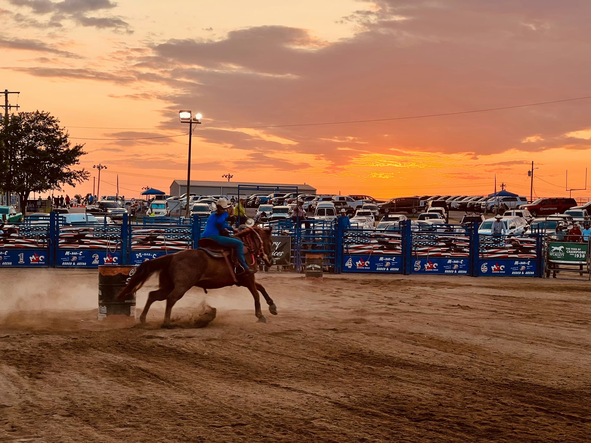 A rodeo arena at sunset, a rider working in the dirt as the sky goes orange behind the stands